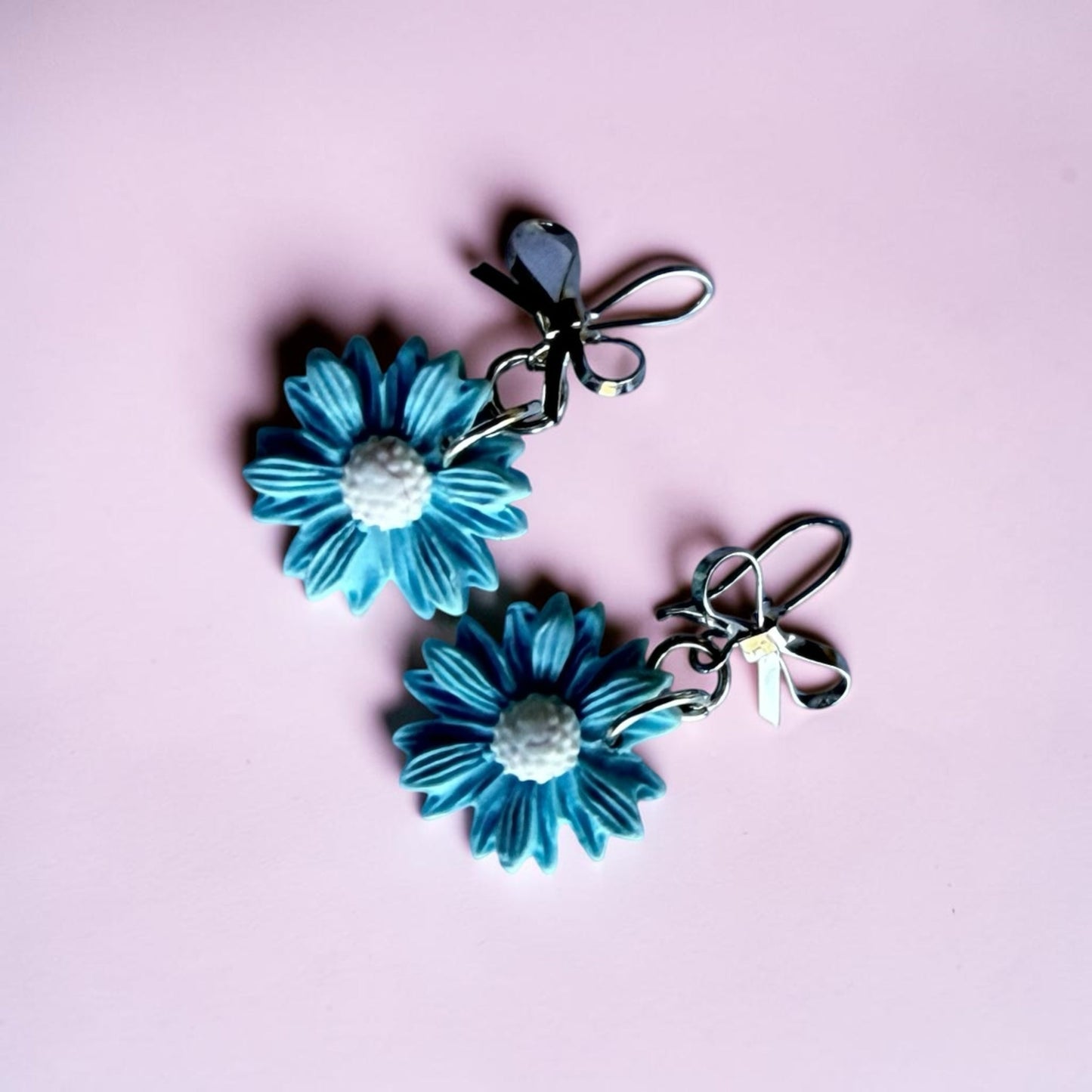 Blue Bottle Crafts handcrafted blue polymer clay flower earrings with silver bow accents on a soft pink background.